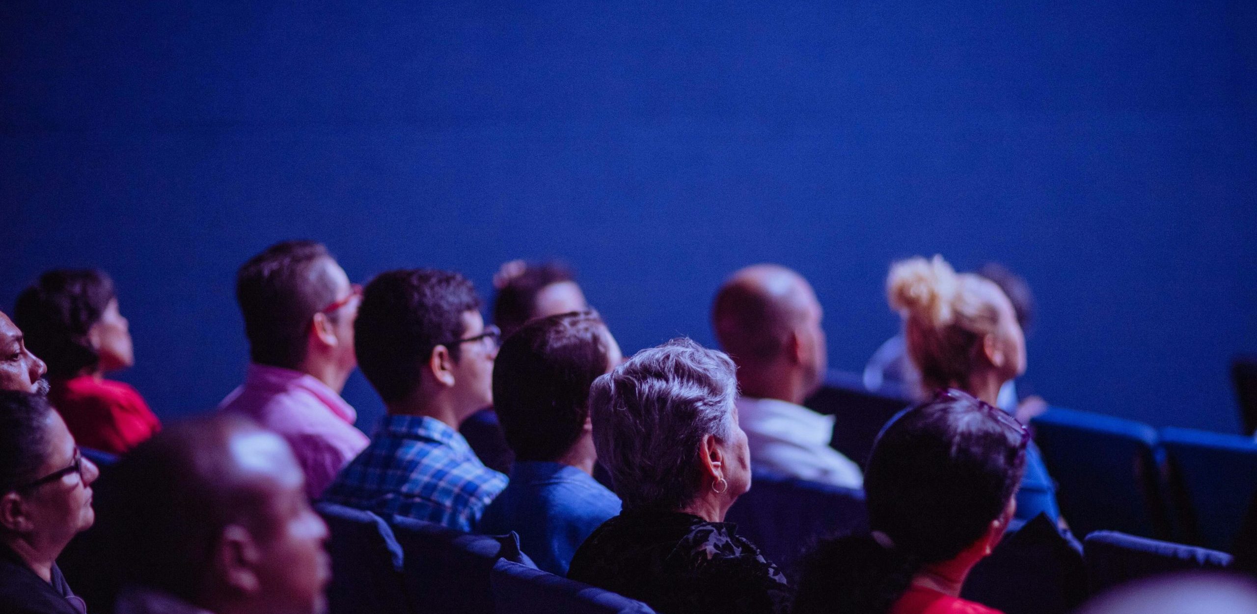 Gros plan sur plusieurs personnes assises en rangées, vues de profil et de dos, attentives dans une salle plongée dans une lumière bleutée, comme lors d’une conférence ou d’une projection.