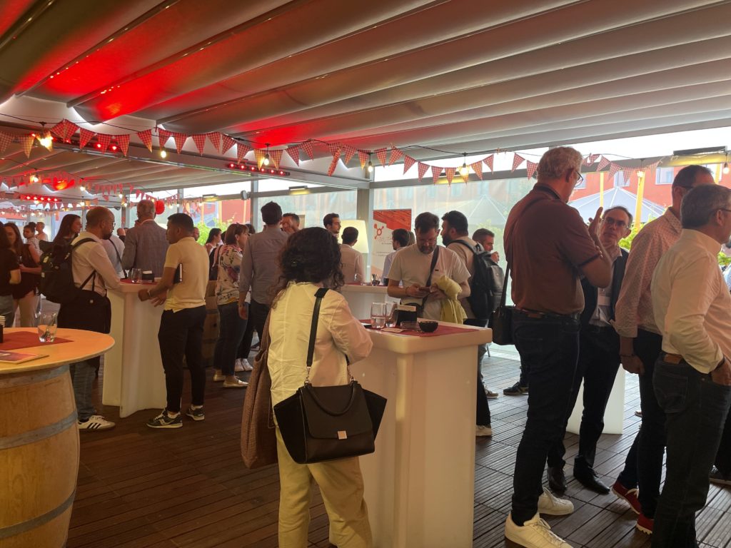 Participants au cocktail networking du roadshow Odoo, debout autour de tables hautes et de tonneaux sous une tonnelle décorée de guirlandes rouges.