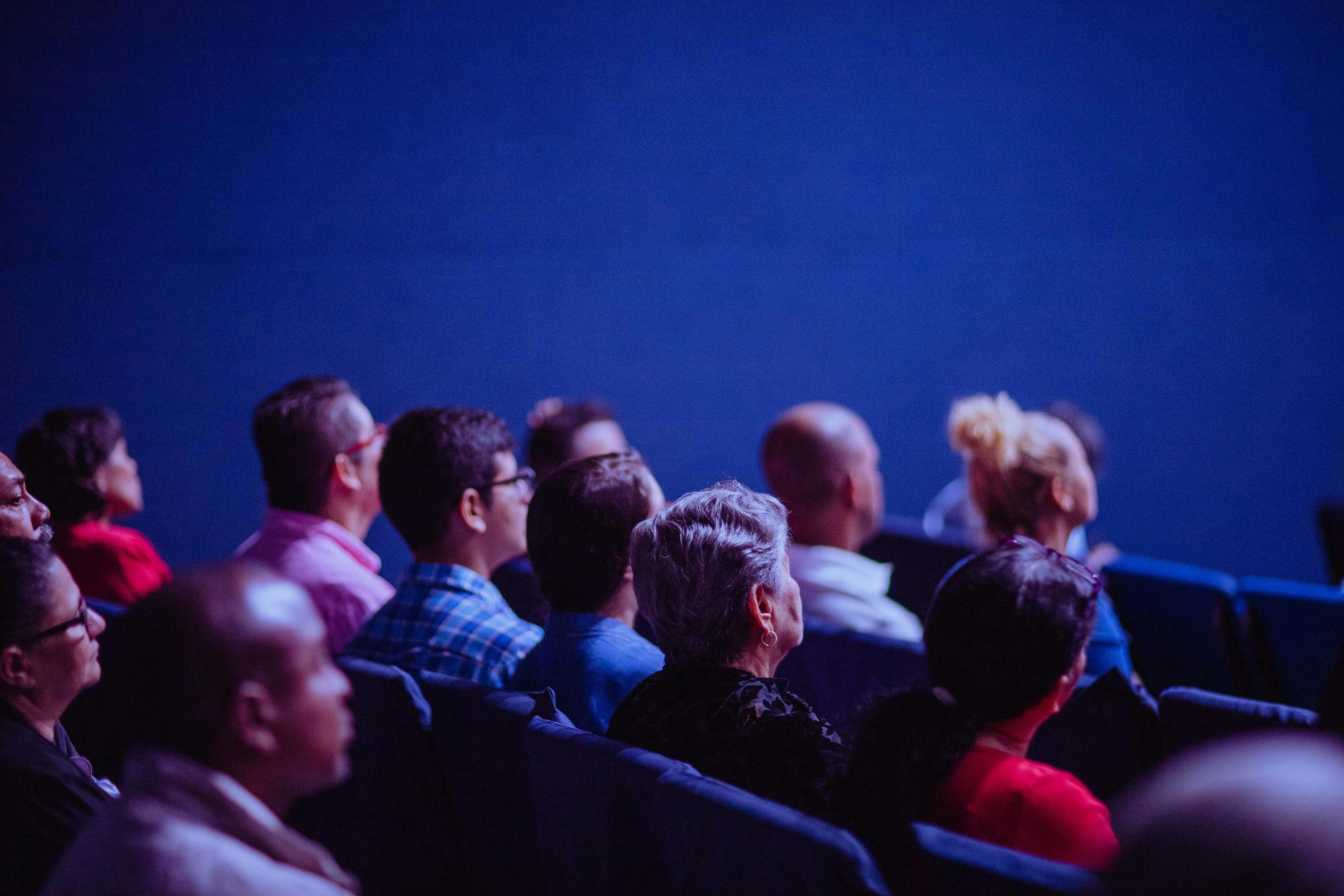 Gros plan sur plusieurs personnes assises en rangées, vues de profil et de dos, attentives dans une salle plongée dans une lumière bleutée, comme lors d’une conférence ou d’une projection.
